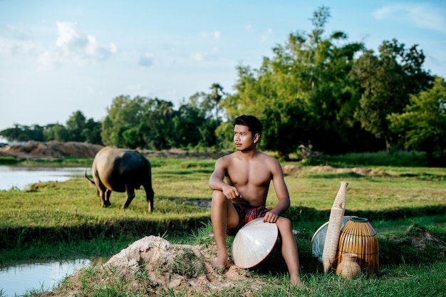 Портрет молодого человека топлес в набедренной повязке в сельской местности, сидя с бамбуковой рыболовной ловушкой