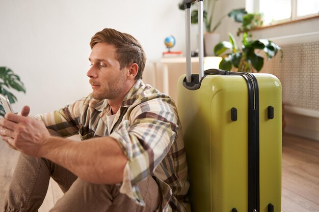 Portrait of young man traveller sits with suitcase and mobile phone chats on smartphone