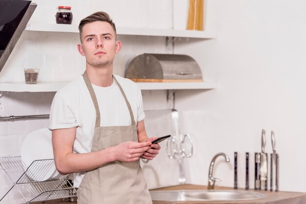 Portrait of a young man wearing apron holding mobile phone in hand looking away