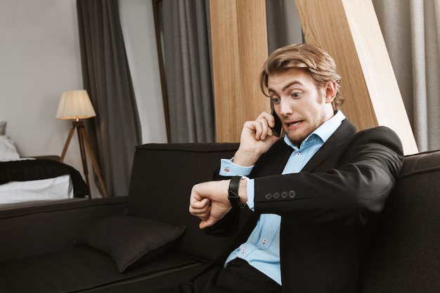 Portrait of young man with blond hair and beard in black suit looking at hand watch with frightened expression being late for meeting with company director