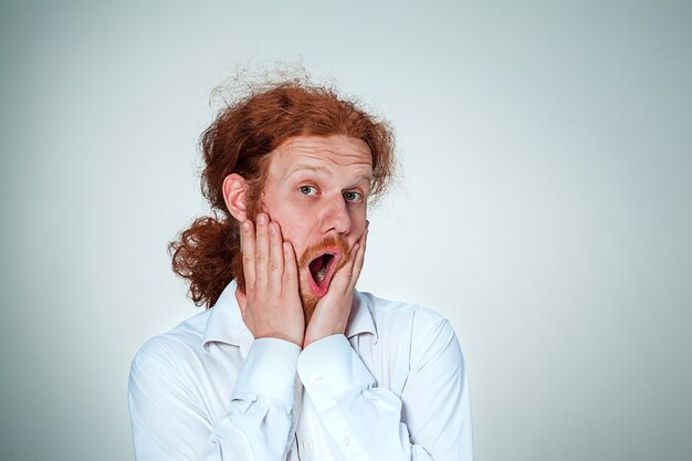 Portrait of young man with long red hair and with shocked facial expression on gray background