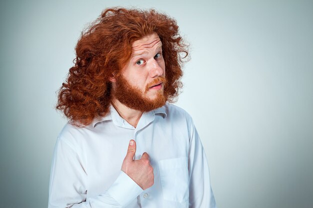 Portrait of young man with long red hair and with shocked facial expression on gray pointing to itself