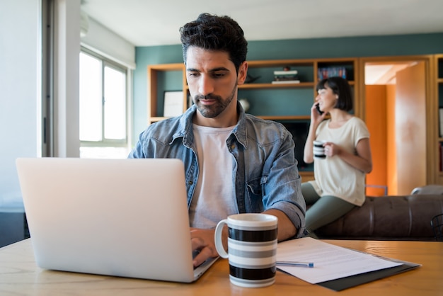 Портрет молодого человека, работающего с ноутбуком из дома, пока женщина разговаривает по телефону на заднем плане