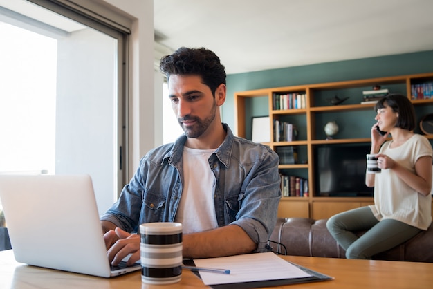 Free photo portrait of young man working with a laptop from home while woman talking on phone at background