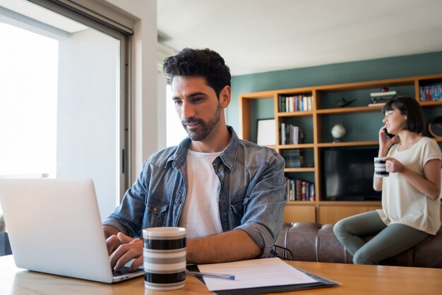 여자가 배경에서 전화로 이야기하는 동안 집에서 노트북으로 작업하는 젊은 남자의 초상화