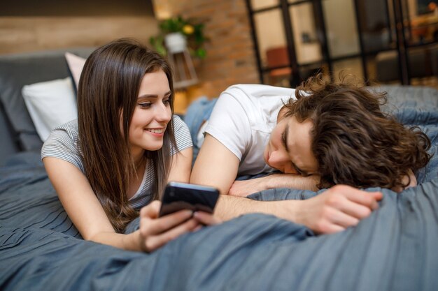 Портрет молодой супружеской пары, лежащей на кровати рано утром, используя смартфон и улыбаясь