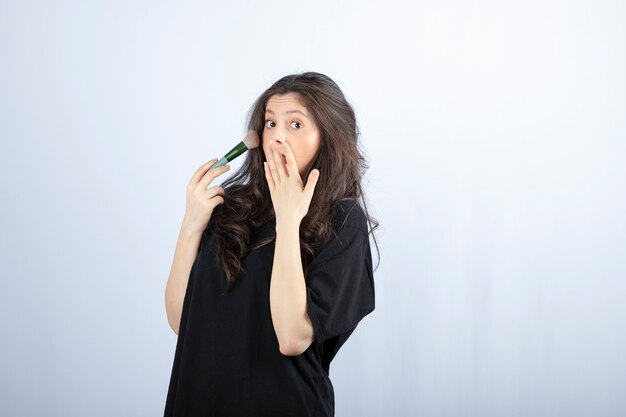 Portrait of young model applying makeup with brush on white wall. 