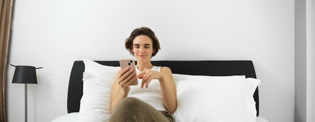 Free photo portrait of young modern woman lying in her bed and using mobile phone resting in bedroom and