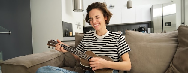 Free photo portrait of young modern woman student playing ukulele at home sitting with small guitar singing and