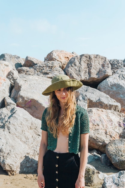 ビーチの岩の近くに立っている彼女の頭の上の帽子を持つ現代の若い女性の肖像画
