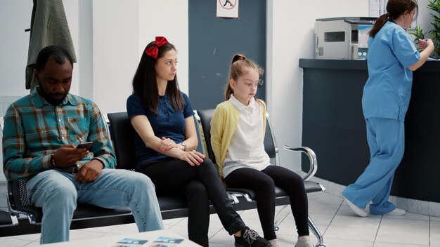 Portrait of young mother with little girl sitting in hospital reception lobby to attend medical consultation with appointment. Waiting room area in healthcare clinic for checkup examination.