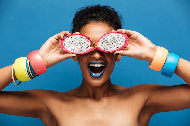 Portrait of young mulatto woman with bracelets on her arms having fun covering eyes with ripe pitahaya fruit cut in half, isolated  over blue