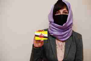 Free photo portrait of young muslim woman wearing formal wear protect face mask and hijab head scarf hold brunei flag card against isolated background coronavirus country concept