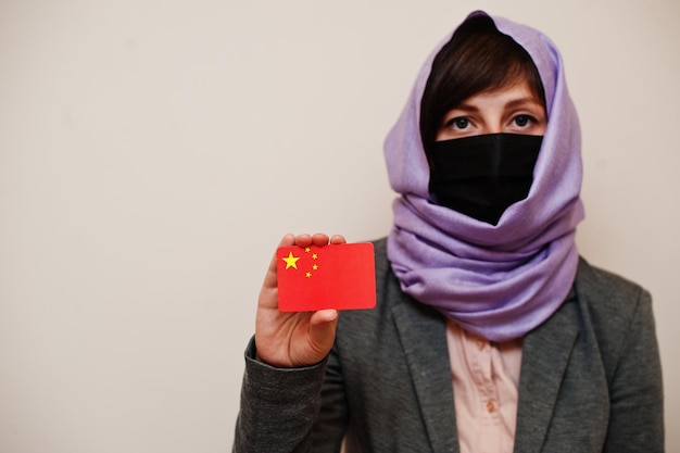 Portrait of young muslim woman wearing formal wear protect face mask and hijab head scarf hold China flag card against isolated background Coronavirus country concept