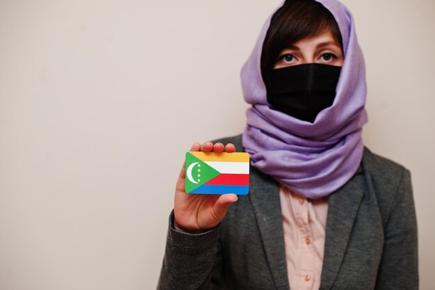 Портрет молодой мусульманки в формальной одежде, защищающей маску для лица и платок в хиджабе, держит карту флага Комор на изолированном фоне Коронавирусная концепция страны