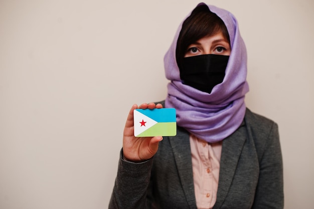 フォーマルな服を着ている若いイスラム教徒の女性の肖像画は、フェイスマスクを保護し、ヒジャーブのヘッドスカーフは、孤立した背景コロナウイルスの国の概念に対してジブチの旗カードを保持します