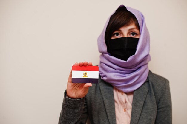 フォーマルな服を着て若いイスラム教徒の女性の肖像画は、フェイスマスクを保護し、ヒジャーブのヘッドスカーフは、孤立した背景コロナウイルスの国の概念に対してエジプトの旗カードを保持します