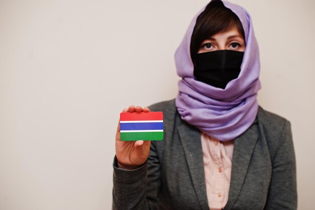 Портрет молодой мусульманки в формальной одежде защищает маску для лица и платок в хиджабе держит карту флага Гамбии на изолированном фоне Коронавирусная концепция страны