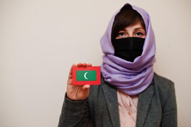 Portrait of young muslim woman wearing formal wear protect face mask and hijab head scarf hold Maldives flag card against isolated background Coronavirus country concept