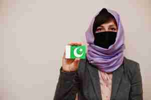 Free photo portrait of young muslim woman wearing formal wear protect face mask and hijab head scarf hold pakistan flag card against isolated background coronavirus country concept