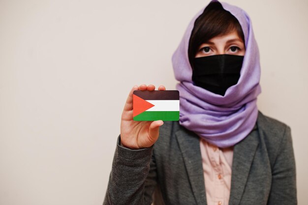 공식적인 복장을 한 젊은 이슬람 여성의 초상화는 얼굴 마스크와 히잡 머리 스카프를 보호하고 고립된 배경 코로나바이러스 국가 개념에 대해 팔레스타인 국기 카드를 들고 있습니다