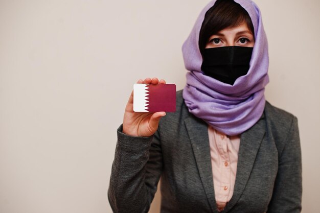Portrait of young muslim woman wearing formal wear protect face mask and hijab head scarf hold Qatar flag card against isolated background Coronavirus country concept