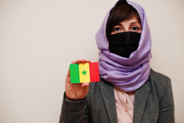 フォーマルな服を着ている若いイスラム教徒の女性の肖像画は、フェイスマスクを保護し、ヒジャーブのヘッドスカーフは、孤立した背景コロナウイルスの国の概念に対してセネガルの旗カードを保持します