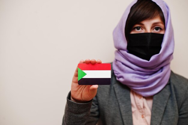 Портрет молодой мусульманки в формальной одежде, защищающей маску для лица и платок в хиджабе, держит карту флага Судана на изолированном фоне Коронавирусная концепция страны