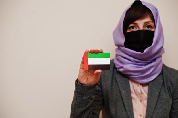 フォーマルな服を着ている若いイスラム教徒の女性の肖像画は、フェイスマスクを保護し、ヒジャーブのヘッドスカーフは、孤立した背景コロナウイルスの国の概念に対してアラブ首長国連邦の旗カードを保持します