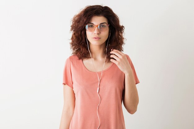 Portrait of young natural hipster pretty woman with curly hairstyle in pink shirt posing wearing glasses isolated on white studio background, listening to music in earphones