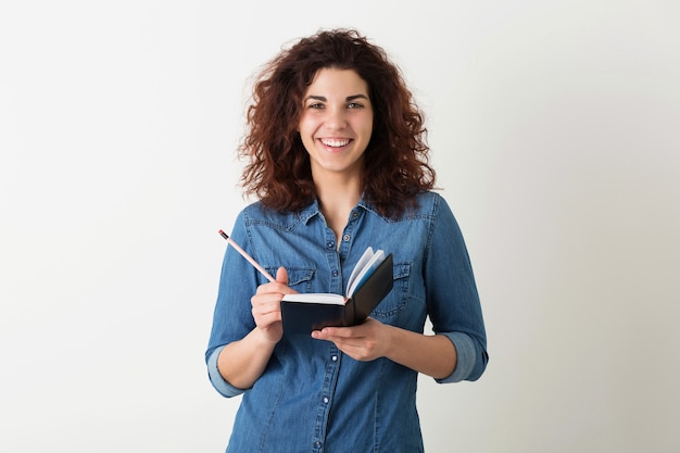 Free photo portrait of young natural hipster smiling pretty woman with curly hairstyle in denim shirt posing with notebook and pen isolated on white studio background, student learning