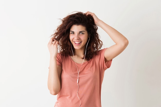 Portrait of young natural looking smiling happy hipster pretty woman with curly hairstyle in pink shirt posing isolated on white studio background, listening to music in earphones