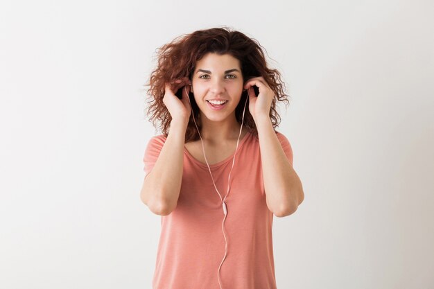Portrait of young natural looking smiling happy hipster pretty woman with curly hairstyle in pink shirt posing isolated on white studio background, listening to music in earphones