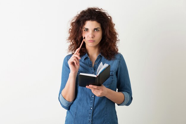 Portrait of young natural smiling pretty woman with curly hairstyle in denim shirt posing with notebook and pen isolated, student learning, thinking on problem