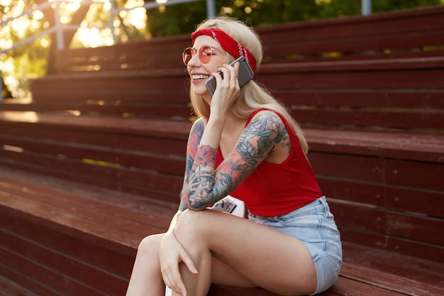Free photo portrait of young nice blonde woman with tattooed arms, holding a smartphone at the ear and talking to his friend. ears in red t-shirt, denim shorts, with knitted bandana on her head, in red glasses.