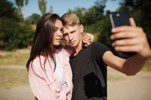 Portrait of young nice couple standing and making selfie in park Cute couple taking photos on cellphone frontal camera