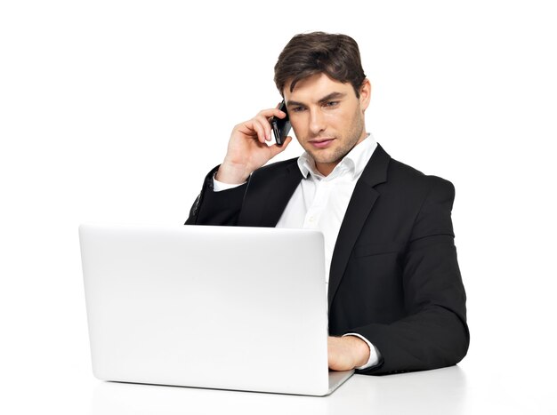 Portrait of young office worker with laptop speaks by mobile phone sitting on table isolated on white.