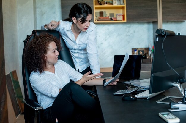 Portrait of young office worker woman sitting at office desk holding documents showing them to colleague with serious expression on face working together in office