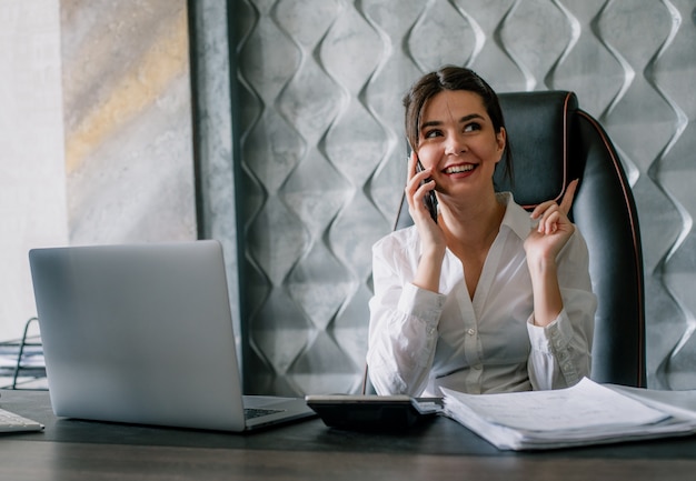 Портрет молодой женщины офисного работника, сидящей за офисным столом, разговаривает по мобильному телефону, улыбаясь со счастливым лицом, рабочий процесс в офисе