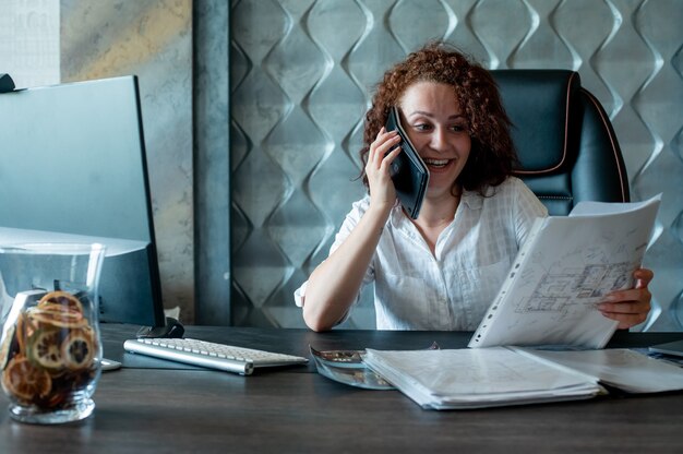 Портрет молодой женщины офисного работника, сидящей за офисным столом, используя калькулятор как мобильный телефон, весело улыбаясь, весело улыбаясь в офисе