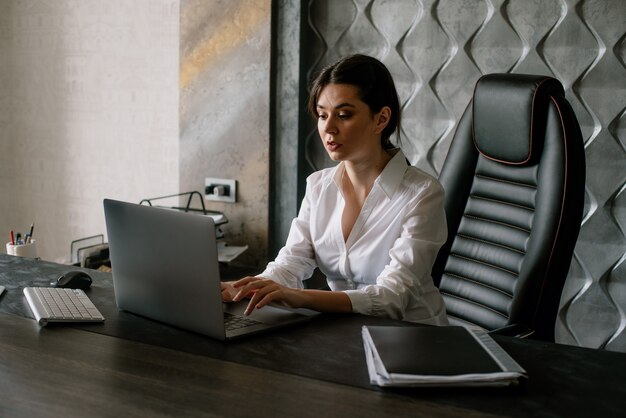 Портрет молодой женщины офисного работника, сидящей за офисным столом, используя портативный компьютер, выглядящей занятой с серьезным и уверенным выражением лица, работающей в офисе