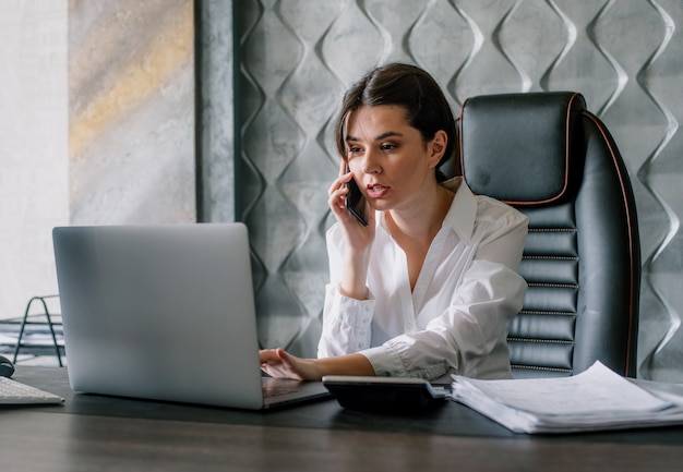 Portrait of young office worker woman sitting at office desk using laptop computer while talking on mobile phone looking anxious working process in office