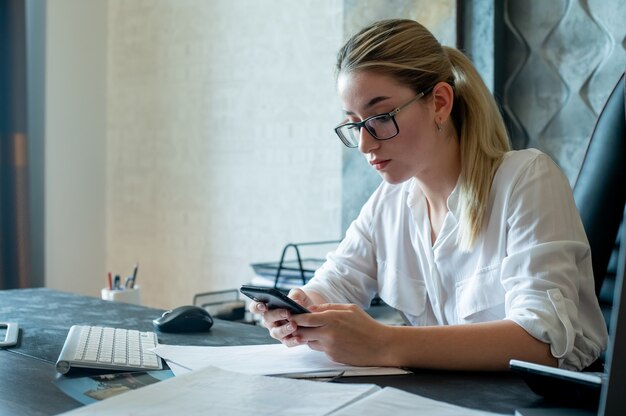 Портрет молодой женщины офисного работника, сидящей за офисным столом с документами, держащими смартфон, смотрящей на экран с серьезным выражением лица, работающей в офисе