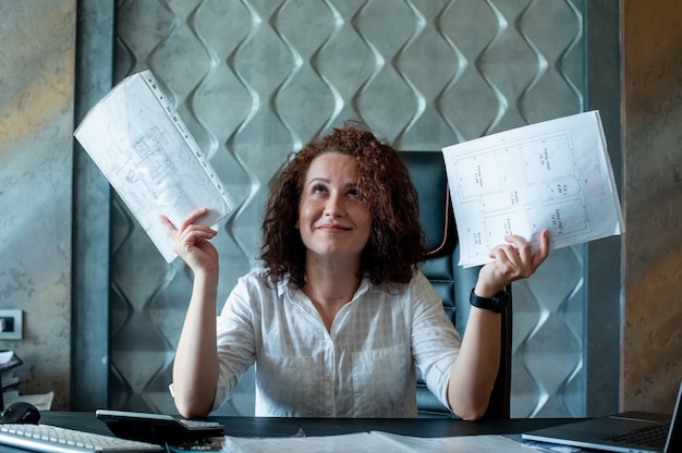 Portrait of young office worker woman sitting at office desk with documents looking up raising hands smiling looking confused working in office