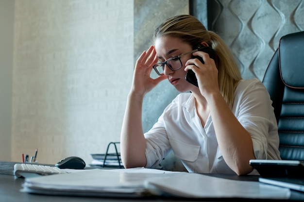 Портрет молодой женщины офисного работника, сидящей за офисным столом с документами, разговаривает по мобильному телефону, нервничает и подчеркивает работу в офисе