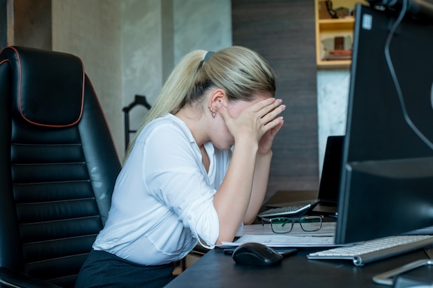 Портрет молодой женщины офисного работника, сидящей за офисным столом с документами, с помощью компьютера, выглядящей усталой и скучающей с головной болью, работая в офисе