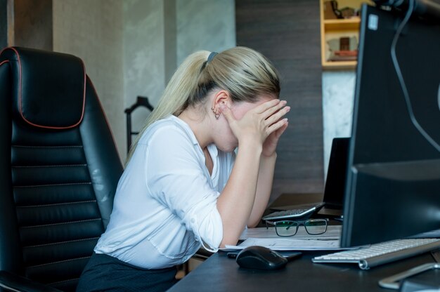 Portrait of young office worker woman sitting at office desk with documents using computer looking tired and bored having headache working in office