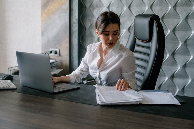 Portrait of young office worker woman sitting at office desk with documents using laptop computer looking confident and busy working in office