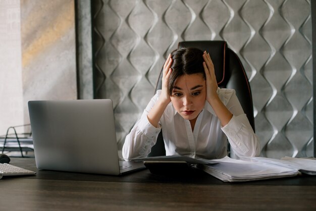 Portrait of young office worker woman sitting at office desk with laptop calculator and documents looking at them nervous and stressed touching her head with arms office concept