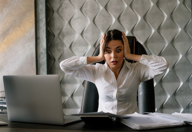 Portrait of young office worker woman sitting at office desk with laptop calculator and documents looking at them shocked touching head office concept